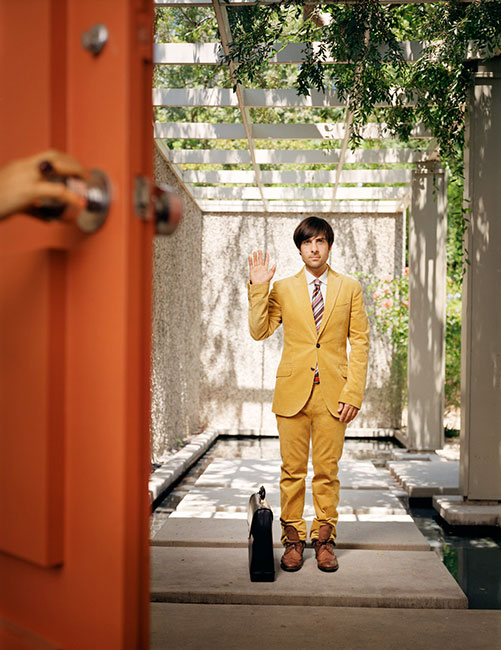 Adult male in yellow suit raises hand near doorway in modern outdoor corridor with plants, concrete, open white trellis roof.