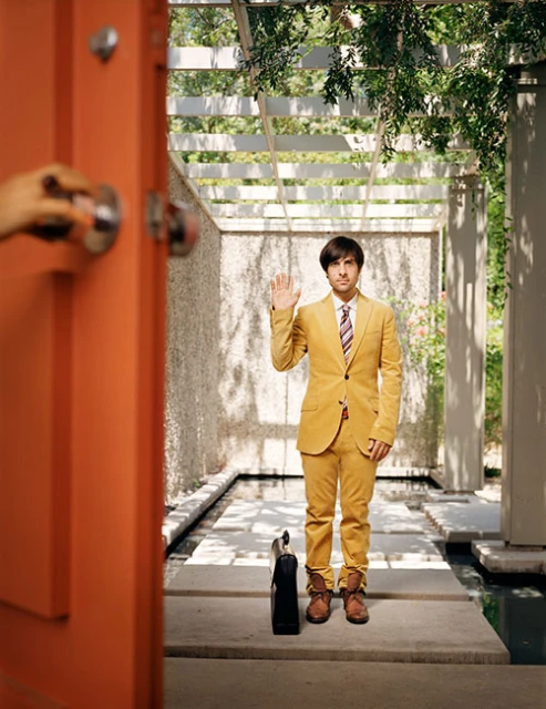 Adult male in yellow suit raises hand near doorway in modern outdoor corridor with plants, concrete, open white trellis roof.