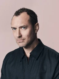 Portrait of actor Jude Law wearing a dark textured zip-up jacket against a neutral pinkish studio backdrop
