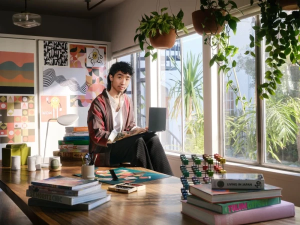 A young man with tattoos sitting on a large wooden table holding a laptop, inside a bright living space filled with books and plants.