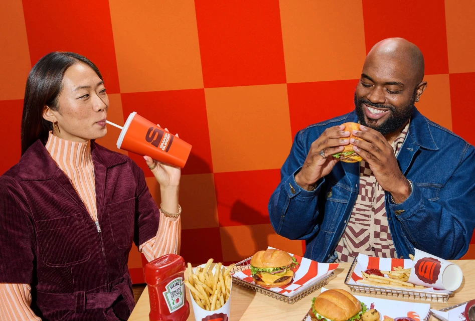 Two people, an Asian woman sipping from a cup and an African American man holding a burger, sit at a table with food.