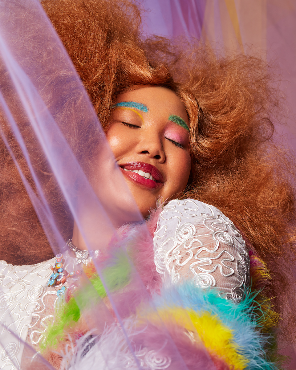 Portrait of a woman with bright makeup and curly ginger hair, wearing a white lace dress against a colorful pastel background.