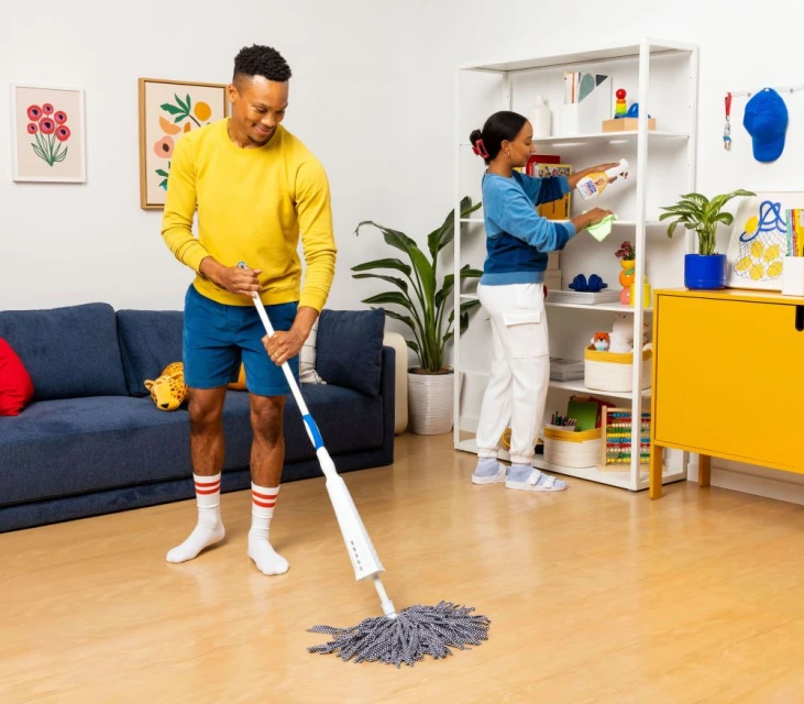 A man uses a mop on the floor while a woman cleans shelves in a bright interior living space, promoting cleaning or home care.