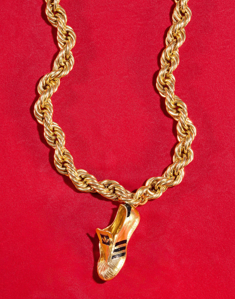 Gold chain necklace with an Adidas shoe pendant. The necklace and pendant are against a red background, shot in a studio setting.