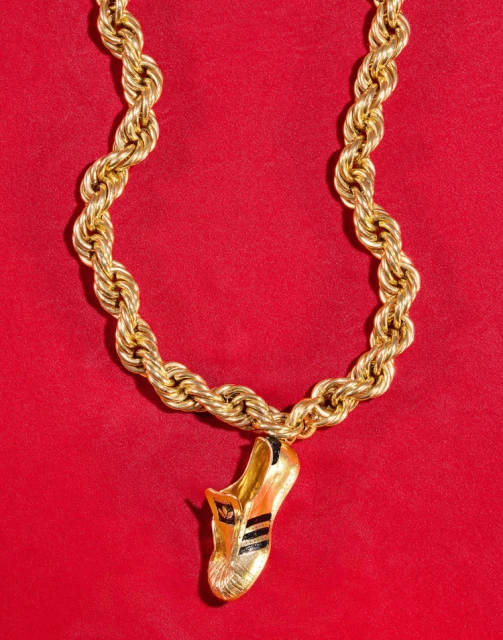 Gold chain necklace with an Adidas shoe pendant. The necklace and pendant are against a red background, shot in a studio setting.