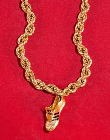 Gold chain necklace with an Adidas shoe pendant. The necklace and pendant are against a red background, shot in a studio setting.
