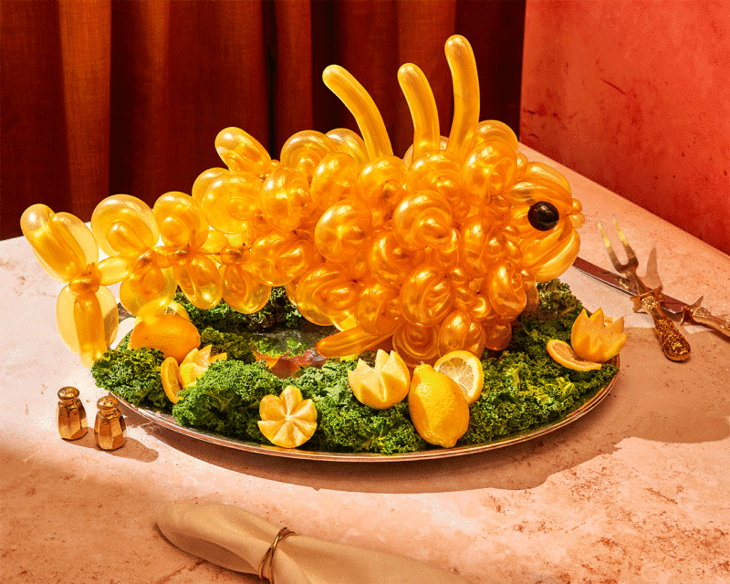 Whimsical yellow balloon-art caterpillar sculpture on a tray with kale and lemons, complemented by salt shakers and antique forks.