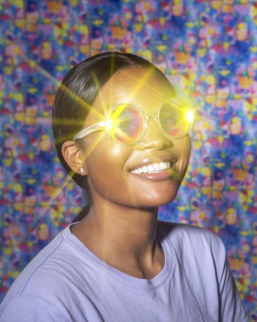 Smiling African-American woman with clear-frame glasses featuring bright light reflections, wearing a purple t-shirt against a patterned backdrop.