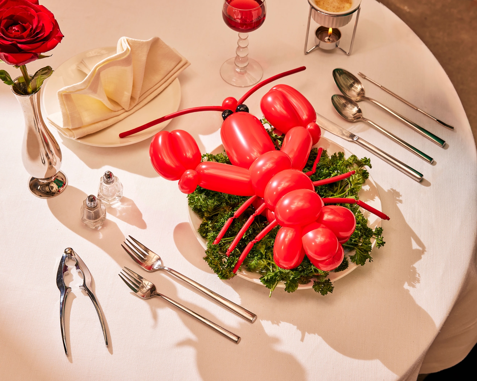 Still life featuring a red balloon lobster centerpiece on a table set with silverware, glassware, and linens, under bright, even lighting.