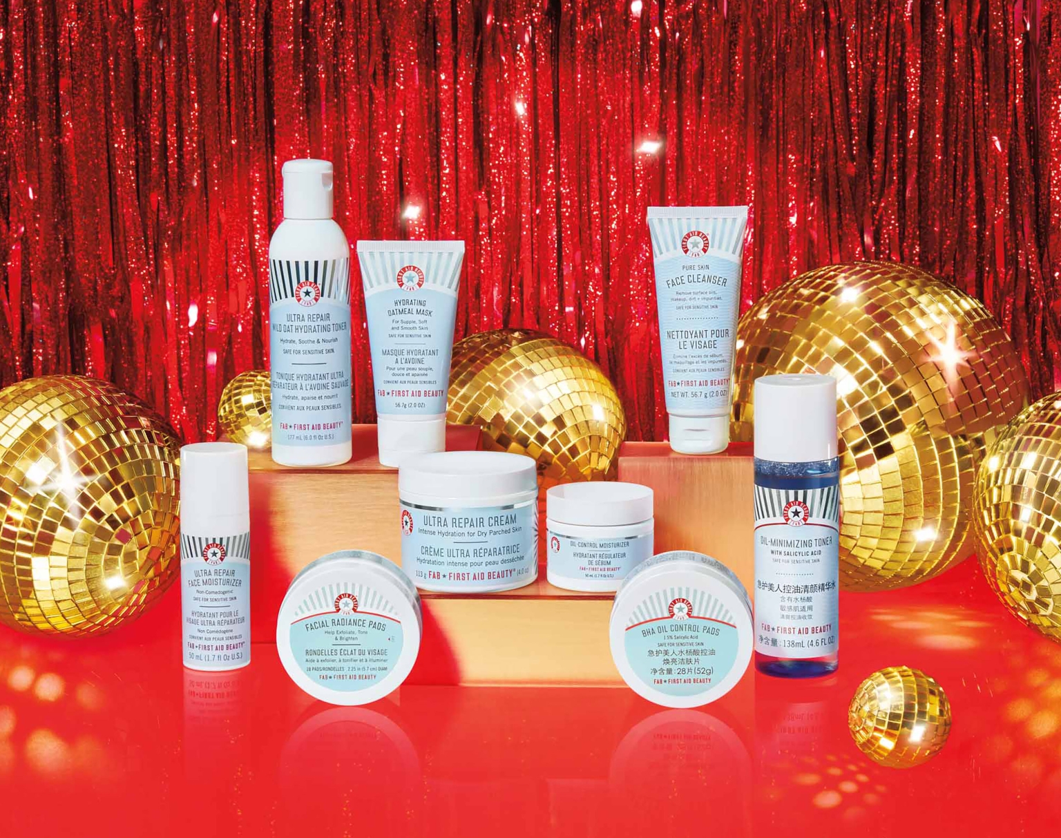 Assortment of First Aid Beauty skincare products including cleansers, creams, and toners are arranged on a red surface with disco ball accents.