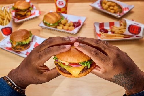 Close-up of a hamburger held by a person, surrounded by french fries and other burgers on a wooden table, fast food concept.
