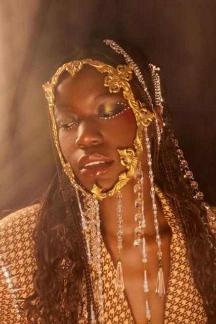 Portrait of a beautiful Black woman with artistic golden face adornments, decorative makeup, and wearing a patterned tan blazer.