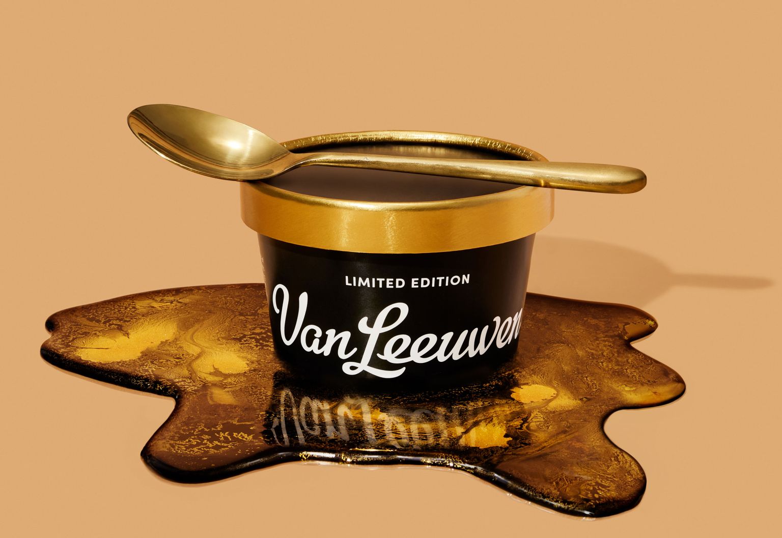 A black pint of Van Leeuwen ice cream with a gold lid and spoon sits atop spilled golden ice cream against a peach background.