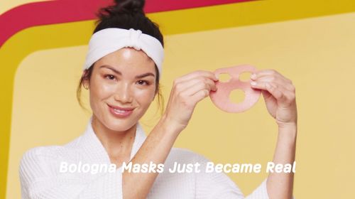 Woman in robe holds bologna face mask with holes for eyes and mouth, posing in front of bright yellow background with retro stripes.