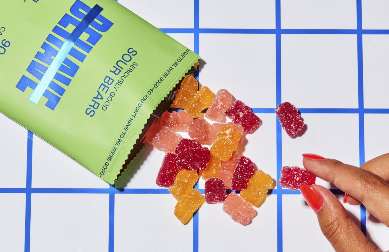 Close-up shot featuring a hand, a vibrant green bag of sour gummy bears, and scattered gummy bears in red, orange, and pink colors.