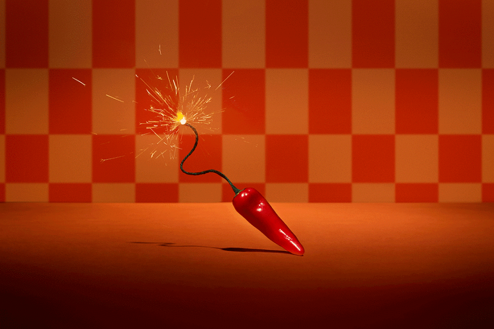 A red chili pepper rests on an orange surface with a checkerboard background, styled as a bomb with a lit wick.