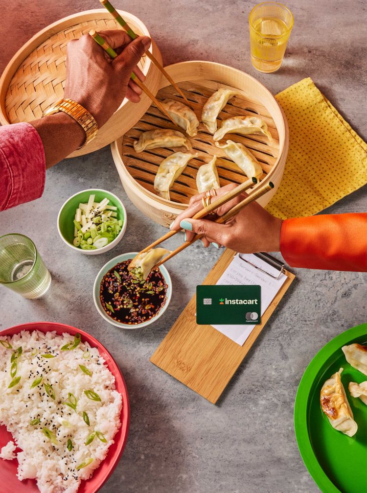 Overhead shot of dumplings in a bamboo steamer and rice on a table, with two people eating with chopsticks; one Instacart card.