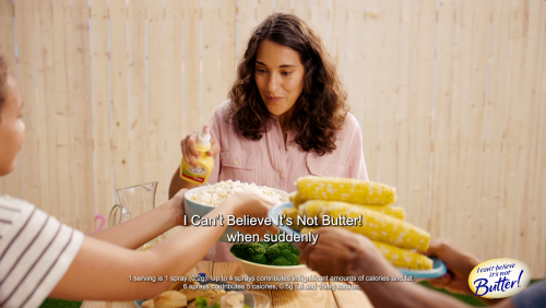 Outdoor picnic scene with a woman spraying 'I Can't Believe It's Not Butter!' on food, surrounded by corn, broccoli, and other edibles.