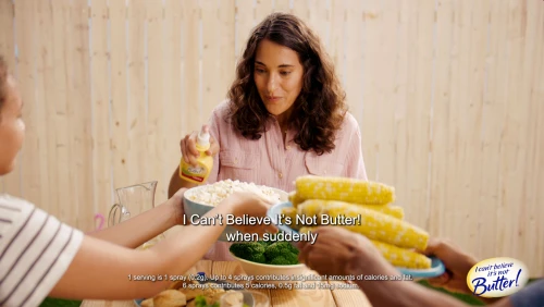 Outdoor picnic scene with a woman spraying 'I Can't Believe It's Not Butter!' on food, surrounded by corn, broccoli, and other edibles.
