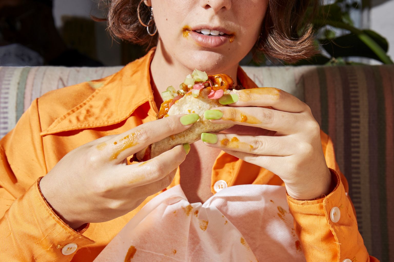 Woman eating messy taco with bright orange shirt, paper bib, and green nail polish, creating a vibrant food-focused image.