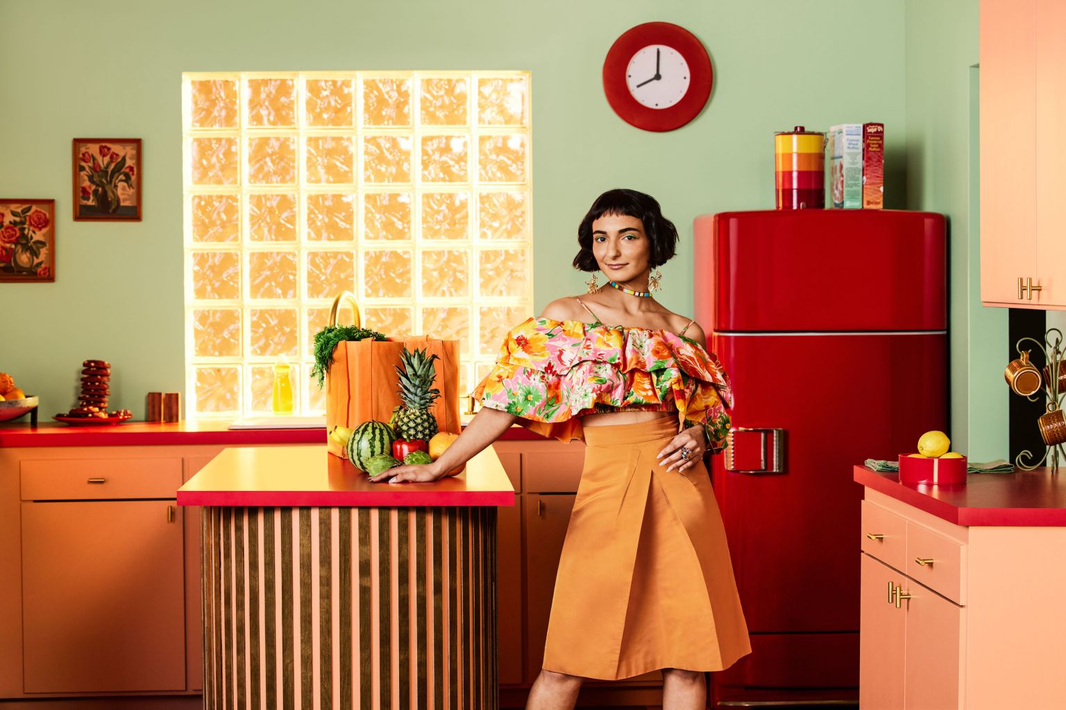 Woman in floral crop top and orange skirt poses in a retro-style kitchen with a red refrigerator and fresh produce on the counter.