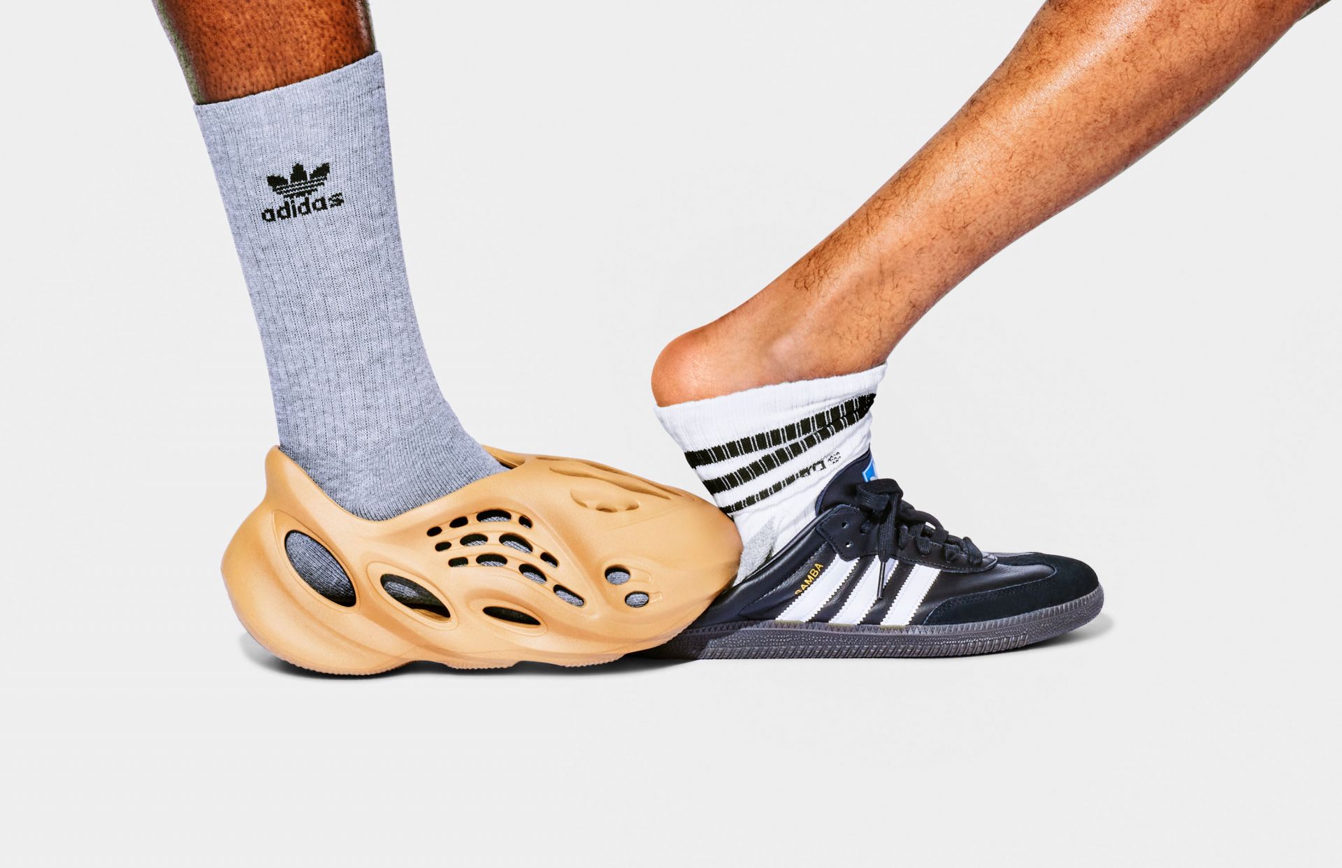 Two feet wear Adidas sneakers and a foam shoe on a white background. One foot wears a gray Adidas sock and the other a striped sock.