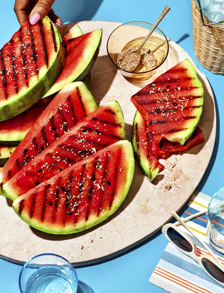 Overhead shot of grilled watermelon slices on a platter with a glass of seasoning, sunglasses, and a striped towel on a blue surface.