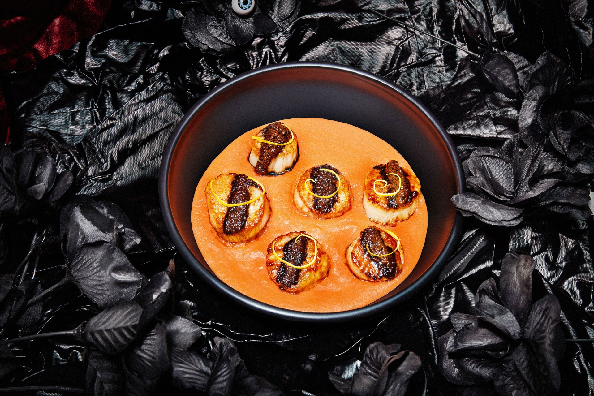 Seared scallops in a black bowl with an orange sauce, garnished with yellow zest, set against a black background of crushed fabric.