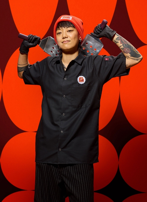 Chef in a black button-down shirt and orange beanie holding two spatulas against a red patterned background