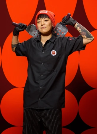 Chef in a black button-down shirt and orange beanie holding two spatulas against a red patterned background