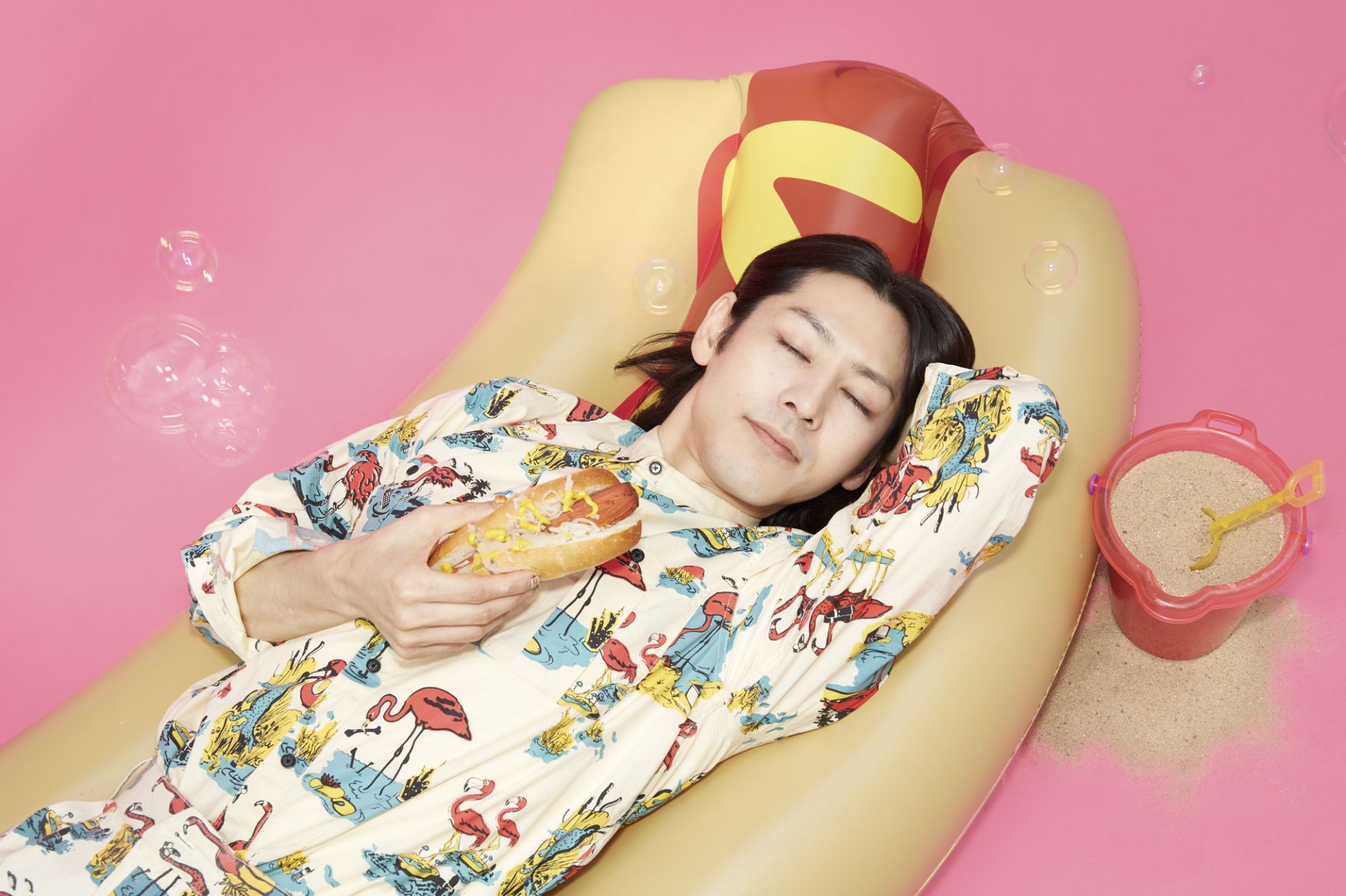 An adult with closed eyes lies on a hotdog floatie holding a hotdog with mustard, with a bucket and spade on a pink background.