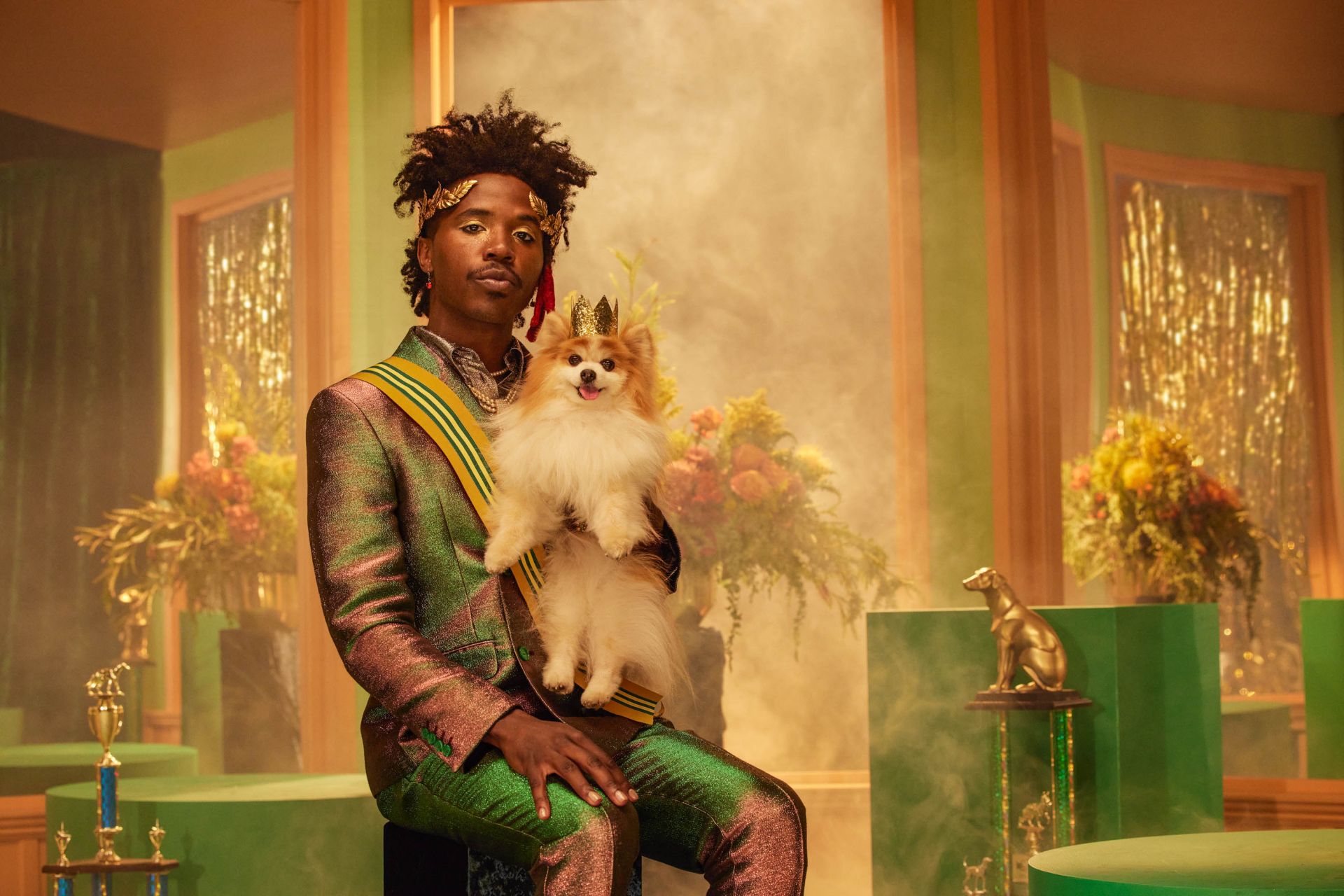 Adult male and dog wearing crowns and sashes in a regal indoor setting with trophies and floral decorations. Ornate, high-fashion style.