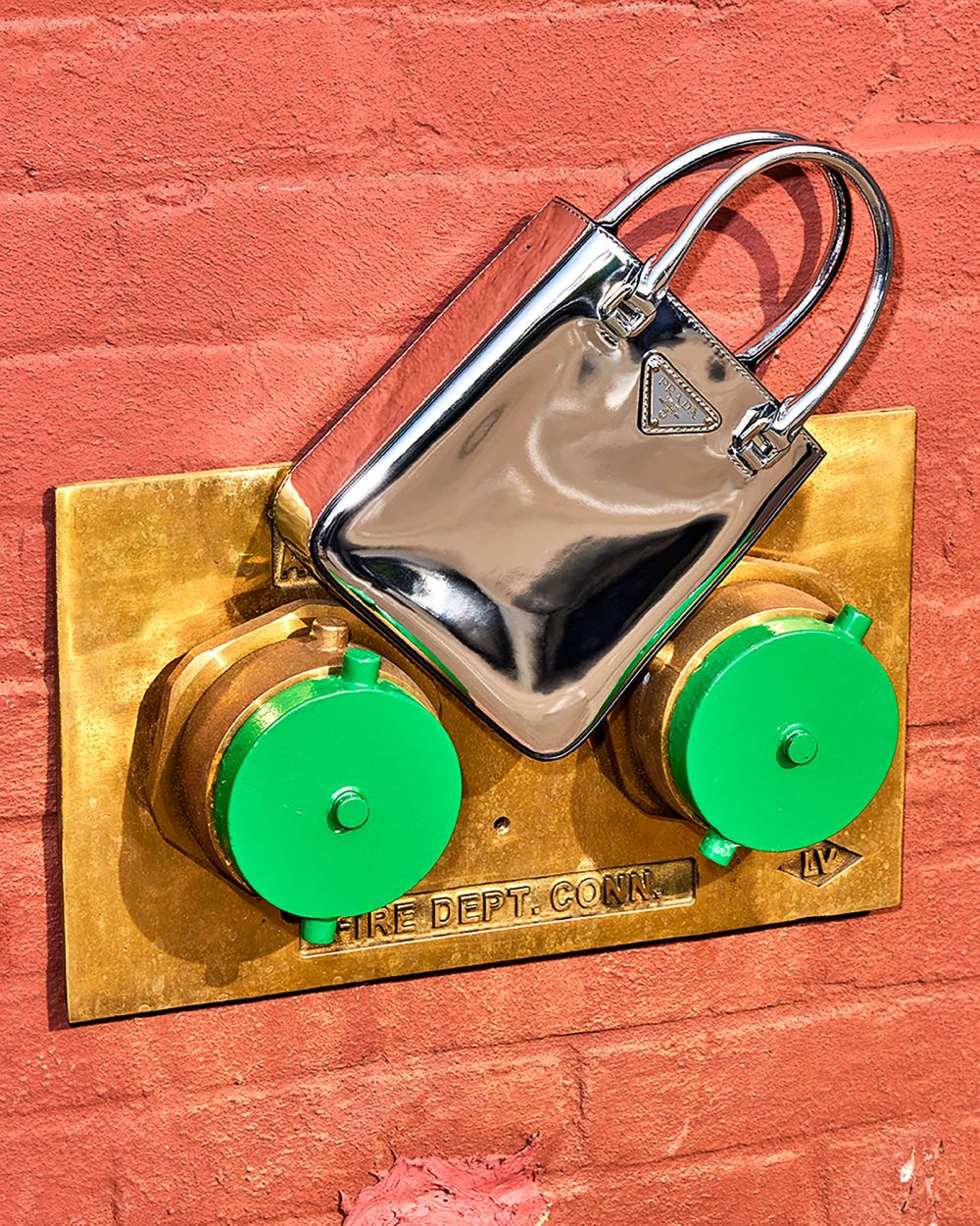 Shiny metallic Prada handbag, silver, leaning against a textured orange brick wall, mounted by a gold fire hydrant fixture with green caps.