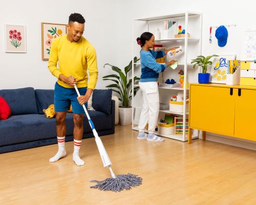 A man mops the floor while a woman organizes a bookshelf in a brightly lit home interior; potted plants and modern decor visible.