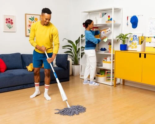 A man mops the floor while a woman organizes a bookshelf in a brightly lit home interior; potted plants and modern decor visible.