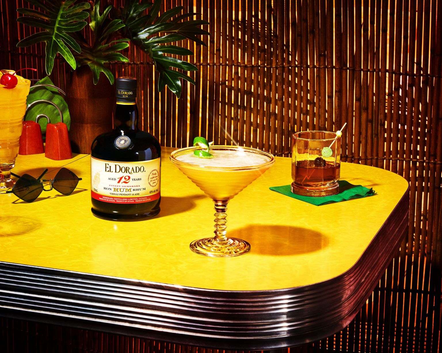 Still life of El Dorado rum bottle and tropical cocktails on a yellow retro bar top against a bamboo wall, travel and food concept.