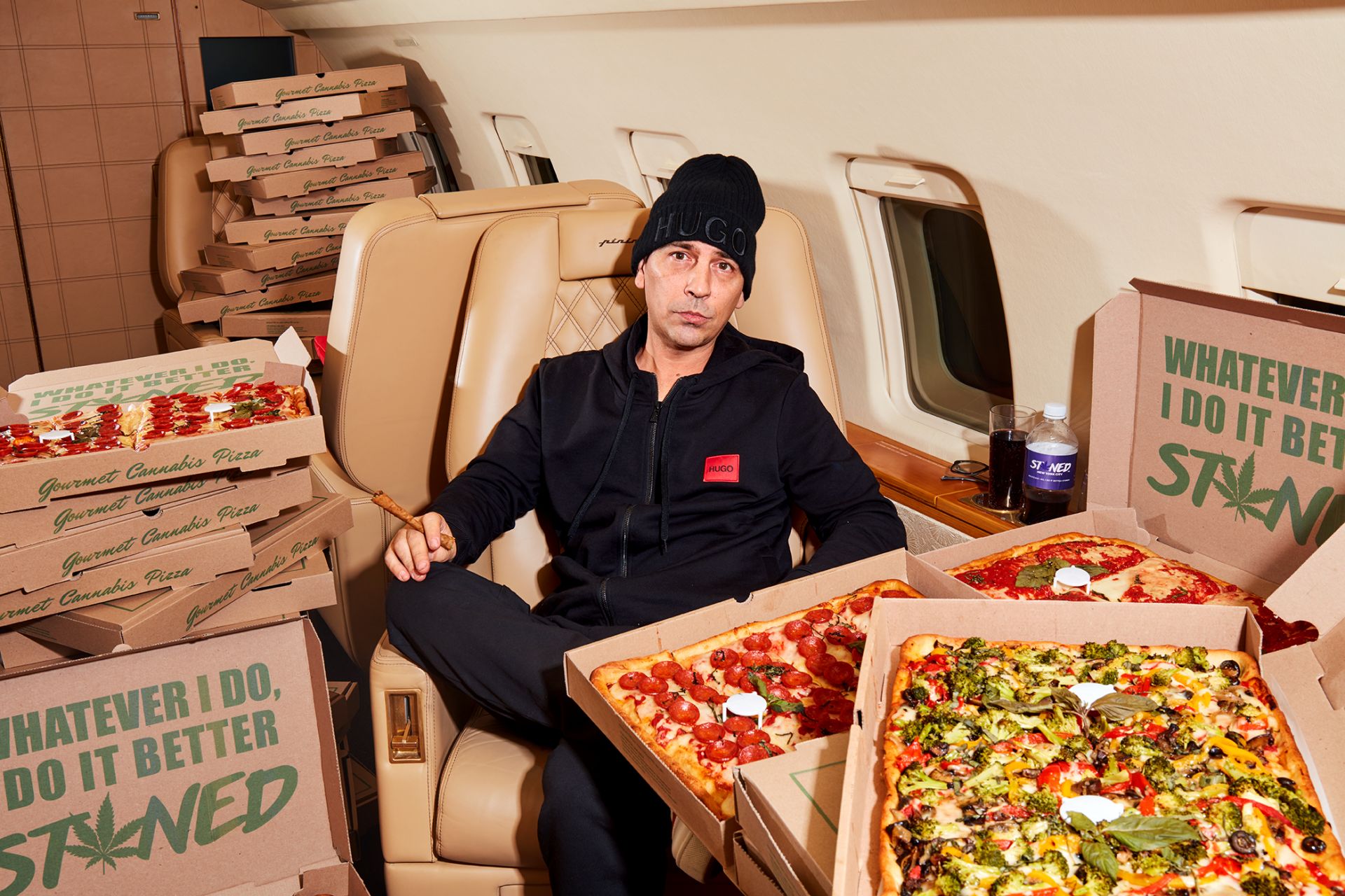Adult male seated in a private jet surrounded by Gourmet Cannabis Pizza boxes, wearing a black Hugo beanie and tracksuit, holding a cigar.