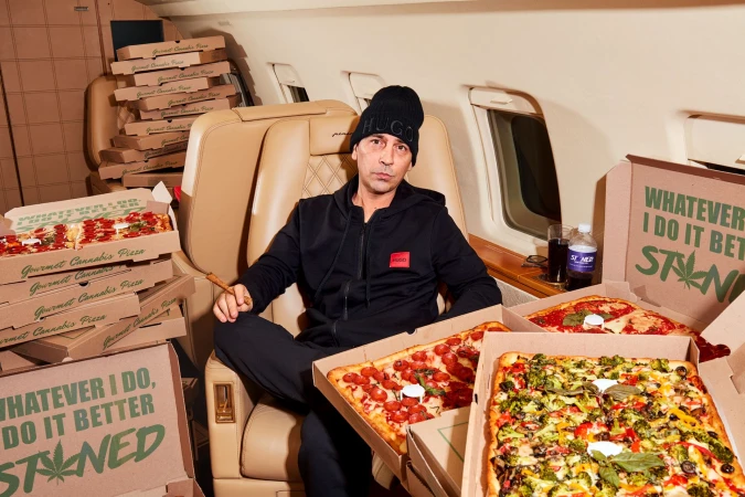 Adult male seated in a private jet surrounded by Gourmet Cannabis Pizza boxes, wearing a black Hugo beanie and tracksuit, holding a cigar.