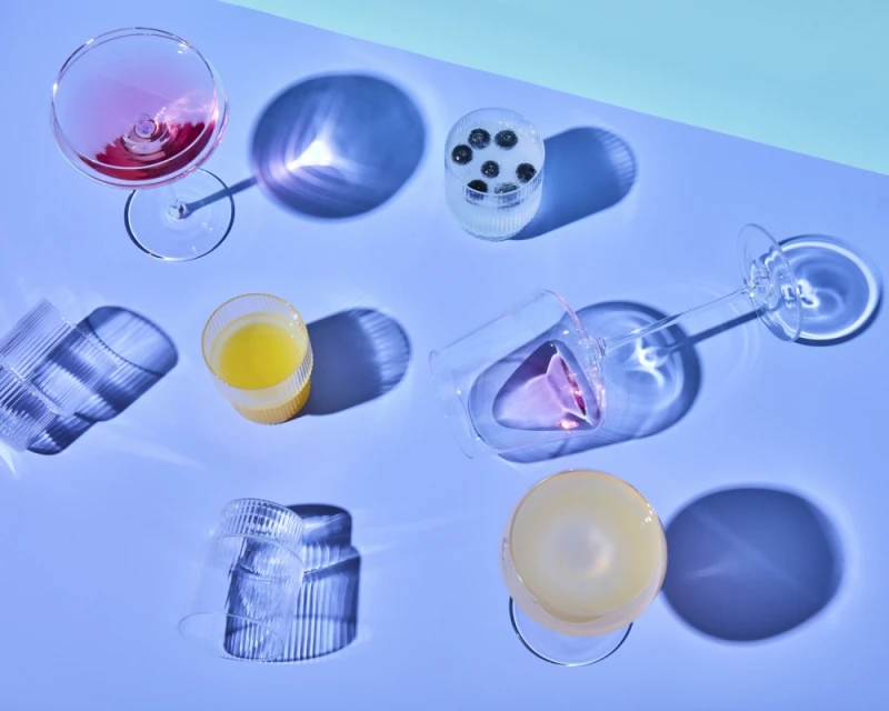 Overhead shot of colorful cocktails and juice in clear textured glasses on a periwinkle background with long shadows and cool lighting.