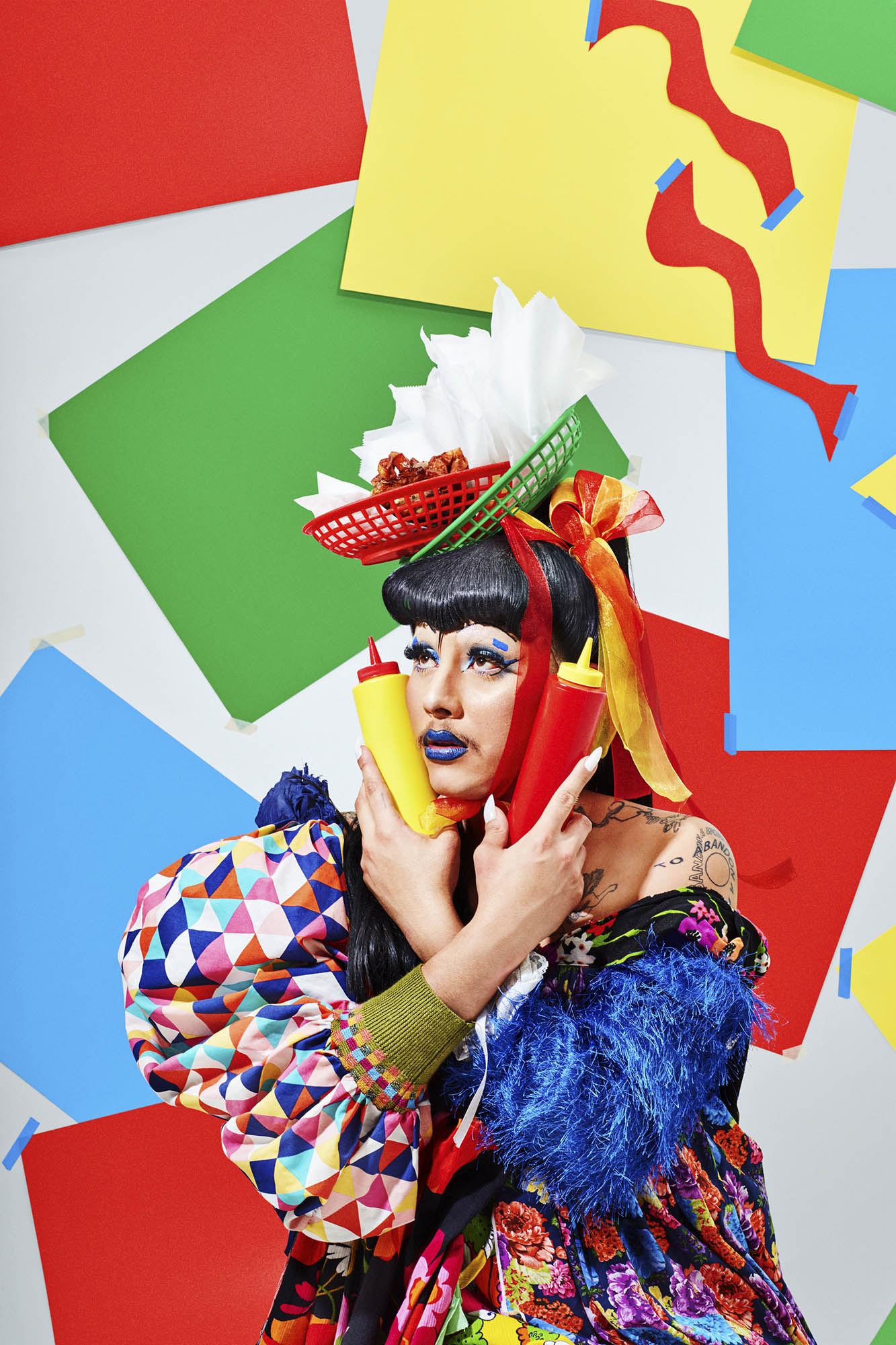 Drag queen with elaborate makeup and headdress poses with colorful background and condiment bottles in a studio setting.