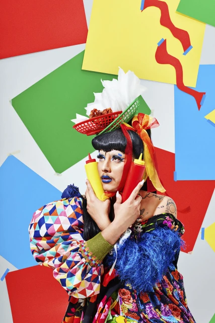 Drag queen with elaborate makeup and headdress poses with colorful background and condiment bottles in a studio setting.