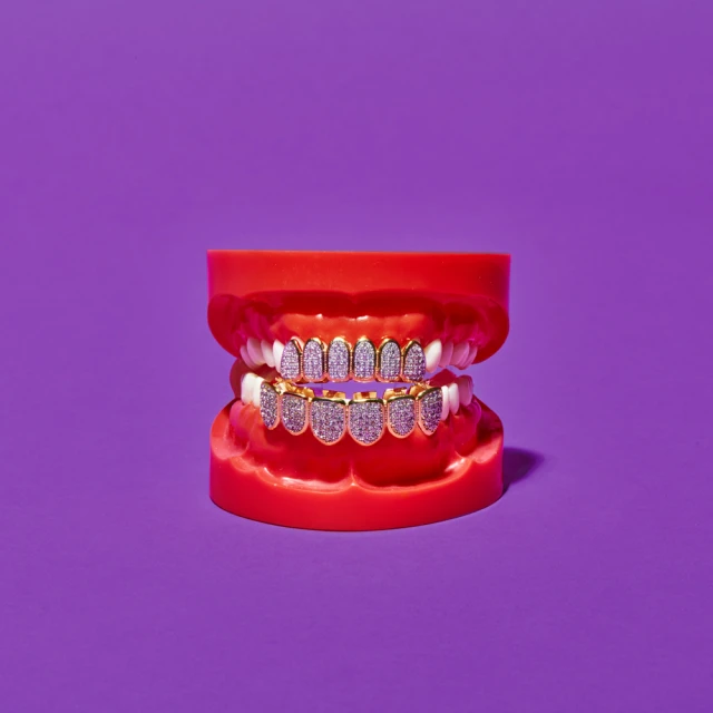 Dental mold displays a gold and jeweled grill, centered on a solid purple background, with simple studio lighting casting soft shadows.