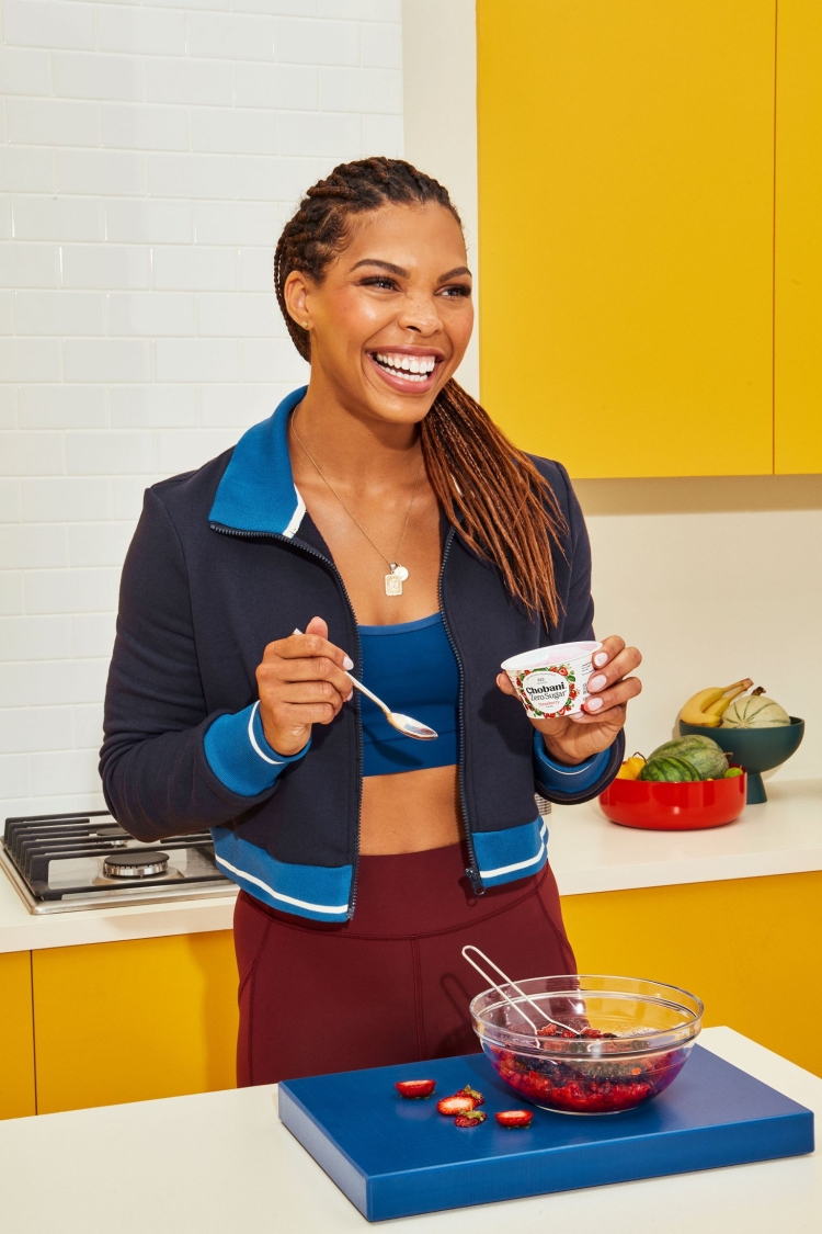 Smiling woman in athletic wear holds a spoonful of berries in a kitchen setting with colorful bowls and countertops, indoors, medium shot.