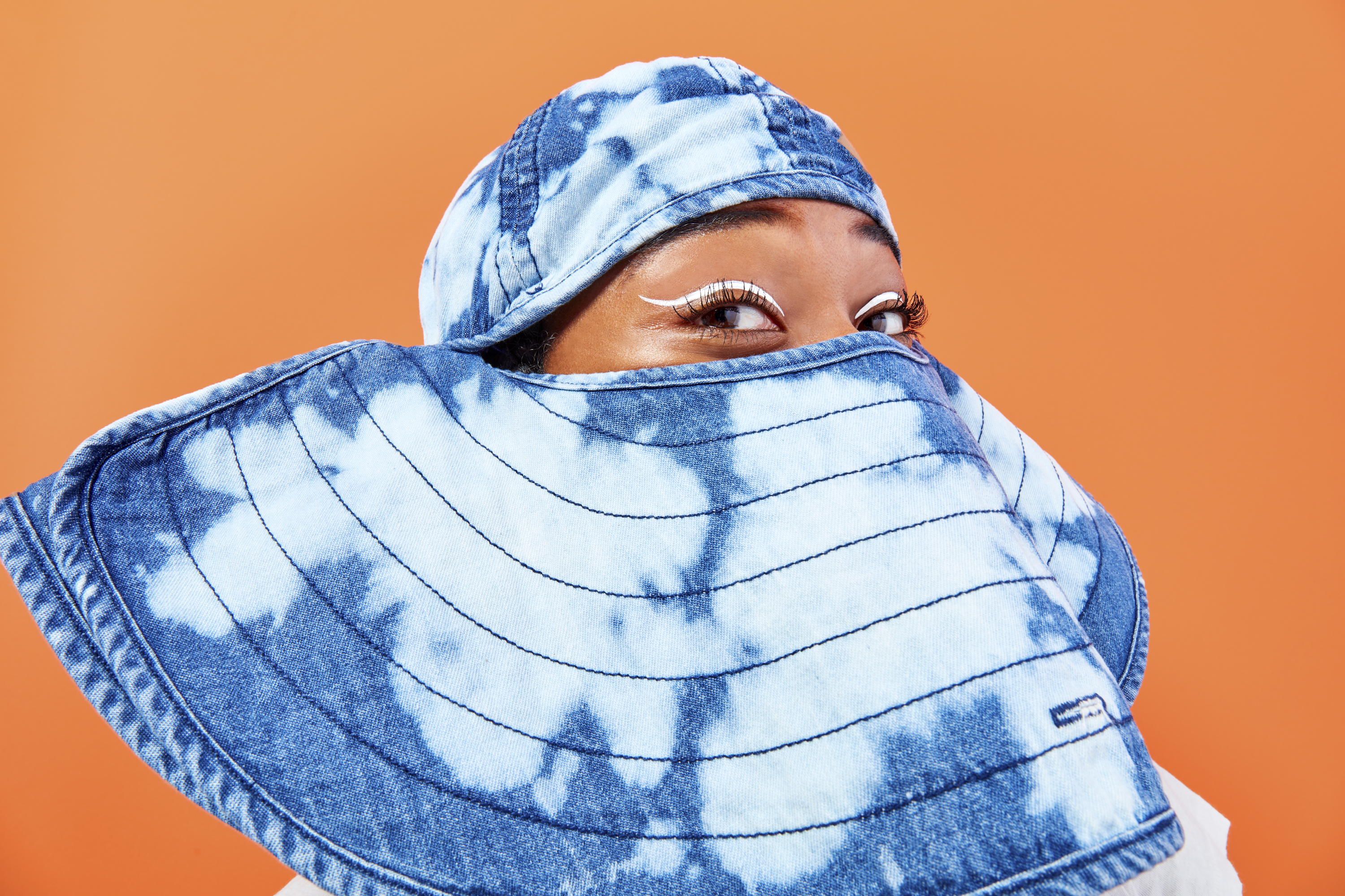 A woman with white eyeliner covers her face with a denim hat with light blue dye patterns on a bright orange background.