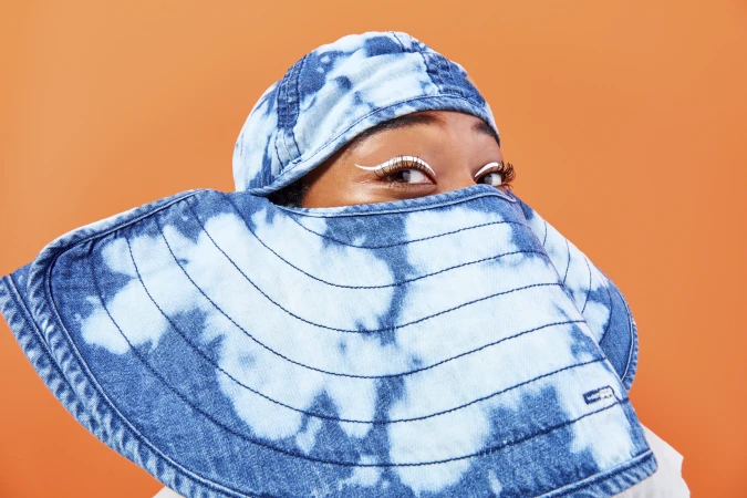 A woman with white eyeliner covers her face with a denim hat with light blue dye patterns on a bright orange background.
