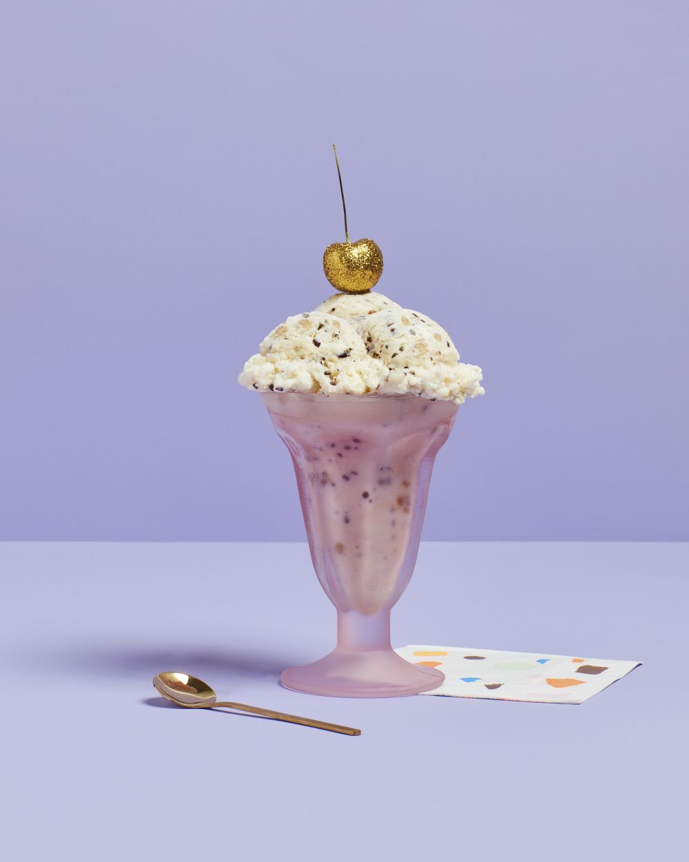Ice cream sundae in a purple glass with a gold cherry on top, accompanied by a gold spoon and a patterned napkin against a purple background.