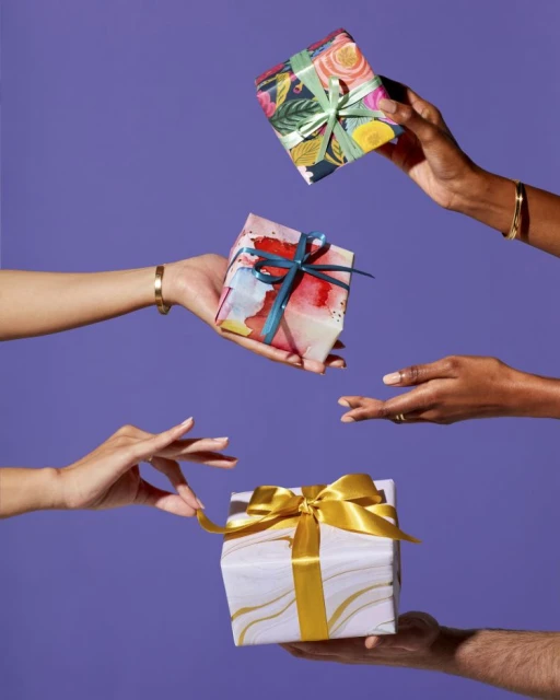 Hands exchange three wrapped gifts with bright ribbons against a vibrant purple background in a celebratory, giving gesture.