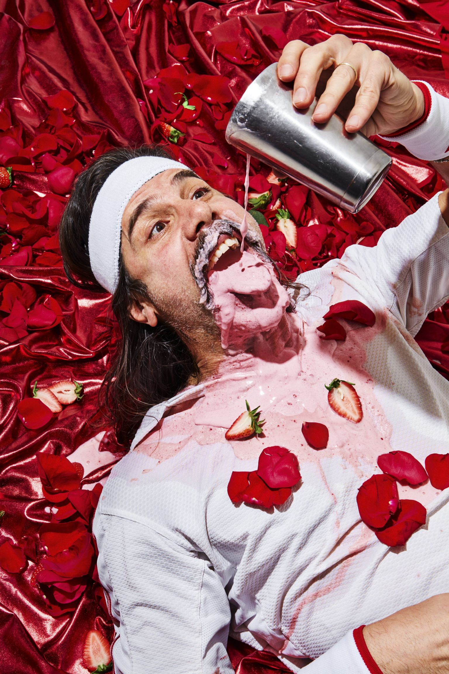 A man lies covered in smoothie and rose petals, face up, on a shiny red surface. He's pouring more of the drink on himself with a shaker.