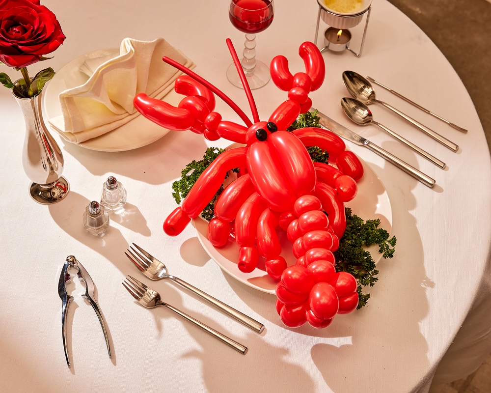 Red balloon lobster centerpiece with silverware, a red rose, and a glass of red liquid on a white tablecloth-covered table.