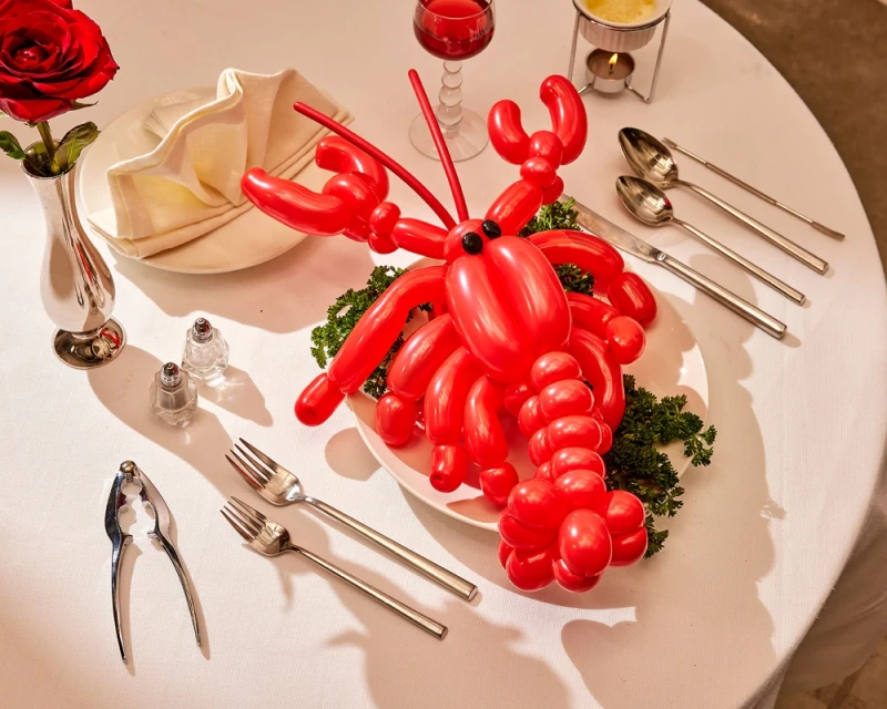 Red balloon lobster centerpiece with silverware, a red rose, and a glass of red liquid on a white tablecloth-covered table.